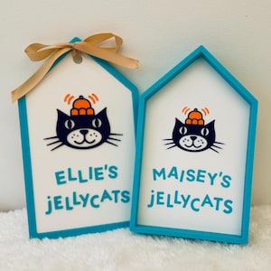 Puede incluir: Dos letreros en forma de casa con marcos color verde azulado y centros blancos. Cada uno presenta una ilustraci&oacute;n de un gato negro con un sombrero naranja y el texto "ELLIE'S JELLYCATS" y "MAISEY'S JELLYCATS" en verde azulado.