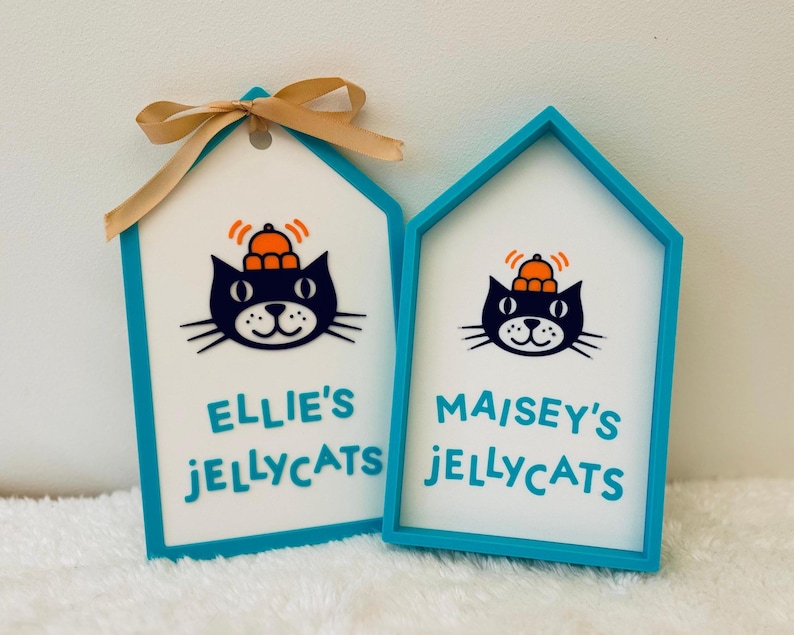 Puede incluir: Dos letreros en forma de casa con marcos color verde azulado y centros blancos. Cada uno presenta una ilustraci&oacute;n de un gato negro con un sombrero naranja y el texto "ELLIE'S JELLYCATS" y "MAISEY'S JELLYCATS" en verde azulado.