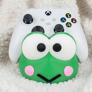 Puede incluir: Un mando de videojuegos inal&aacute;mbrico blanco sobre un soporte verde con forma de rana. El soporte presenta grandes ojos blancos con pupilas negras, mejillas circulares rosas y una boca negra en forma de V. El mando tiene joysticks de color negro.