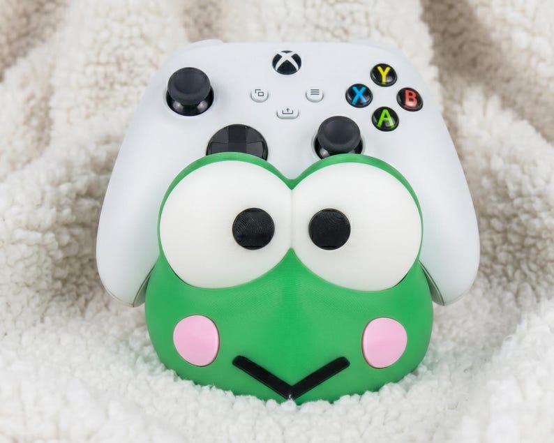 Puede incluir: Un mando de videojuegos inal&aacute;mbrico blanco sobre un soporte verde con forma de rana. El soporte presenta grandes ojos blancos con pupilas negras, mejillas circulares rosas y una boca negra en forma de V. El mando tiene joysticks de color negro.