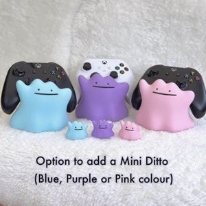 以下が含まれることがあります： ライトブルー、パープル、ピンクのキャラクター「メタモン」の形をしたコントローラースタンド3つ。それぞれゲームコントローラーを保持しています。手前には、お揃いの小さなメタモンのミニフィギュアが3つ置かれています。テキストには「Option to add a Mini Ditto (Blue, Purple or Pink colour)」と書かれています。