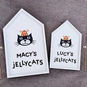 May include: Two white, house-shaped wooden signs. Each sign features a black cat illustration with an orange hat and the words "MACY'S JELLYCATS" and "LUCY'S JELLYCATS". The larger sign is 26.2 cm (10.3 inches) tall, and the smaller sign is 18.8 cm (7.4 inches) tall.