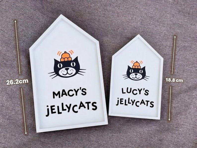 May include: Two white, house-shaped wooden signs. Each sign features a black cat illustration with an orange hat and the words "MACY'S JELLYCATS" and "LUCY'S JELLYCATS". The larger sign is 26.2 cm (10.3 inches) tall, and the smaller sign is 18.8 cm (7.4 inches) tall.