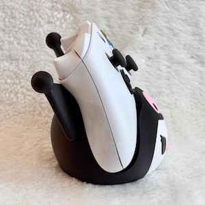 May include: A white video game controller resting in a black and white character-themed stand. The stand features black upright supports with rounded tips and a rounded base with pink and white accents, designed to hold the controller upright.