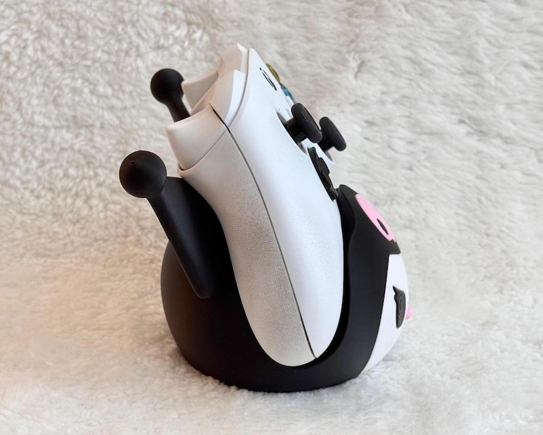 May include: A white video game controller resting in a black and white character-themed stand. The stand features black upright supports with rounded tips and a rounded base with pink and white accents, designed to hold the controller upright.