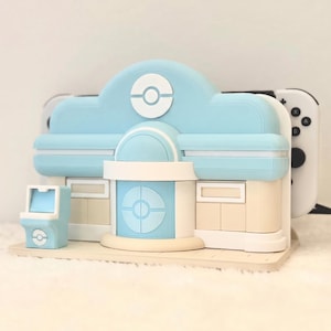 May include: A light blue and beige gaming console dock shaped like a building with a central circular emblem. The structure features a small matching arcade-style side unit and holds a white gaming console with black buttons visible on the right side.