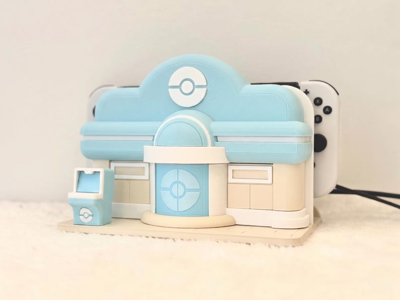 May include: A light blue and beige gaming console dock shaped like a building with a central circular emblem. The structure features a small matching arcade-style side unit and holds a white gaming console with black buttons visible on the right side.