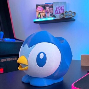 K&ouml;nnte beinhalten: Eine blau-wei&szlig;e, 3D-gedruckte Piplup-Figur, ein Pok&eacute;mon-Charakter, mit gelbem Schnabel. Die Figur steht auf einer schwarzen Oberfl&auml;che. Im Hintergrund zeigt ein Regal Filmh&uuml;llen und Schlagringe.