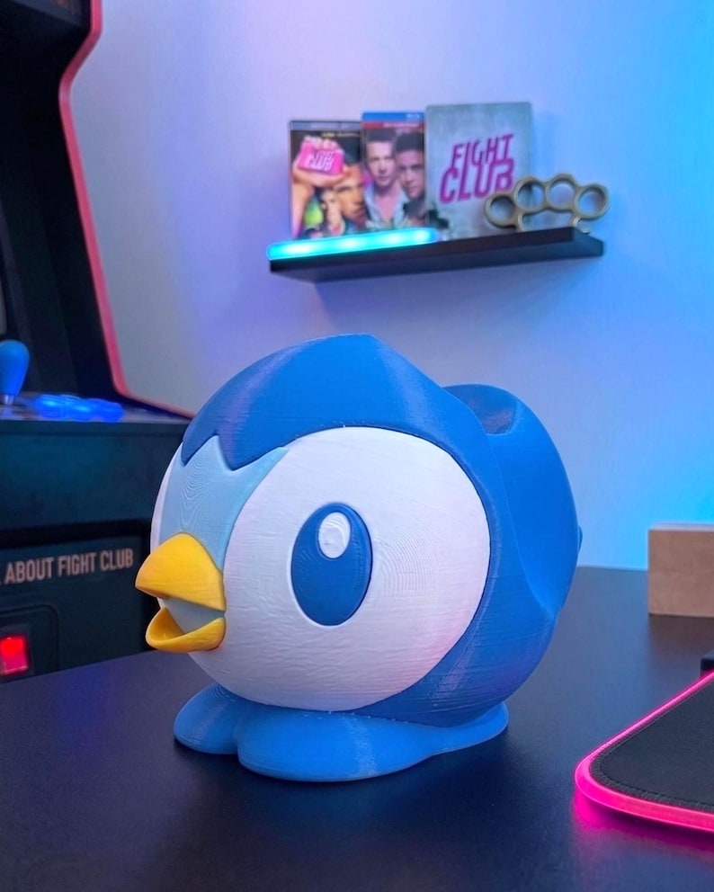 K&ouml;nnte beinhalten: Eine blau-wei&szlig;e, 3D-gedruckte Piplup-Figur, ein Pok&eacute;mon-Charakter, mit gelbem Schnabel. Die Figur steht auf einer schwarzen Oberfl&auml;che. Im Hintergrund zeigt ein Regal Filmh&uuml;llen und Schlagringe.