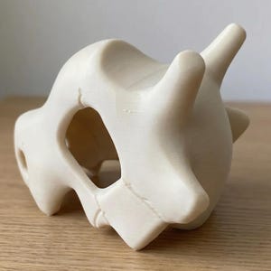 May include: A cream-colored, 3D-printed Cubone skull figurine. The skull features a large eye socket and two prominent horns. The figurine is sitting on a wooden surface.