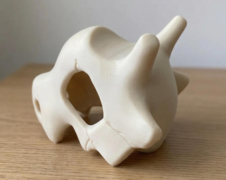 May include: A cream-colored, 3D-printed Cubone skull figurine. The skull features a large eye socket and two prominent horns. The figurine is sitting on a wooden surface.