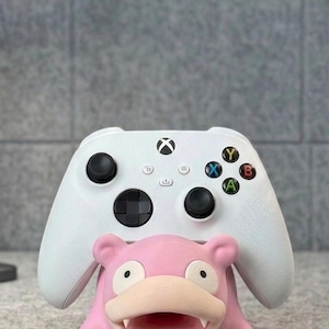Pode incluir: Um controle Xbox branco repousa sobre um suporte rosa em forma de Slowpoke. O controle tem joysticks pretos e bot&otilde;es coloridos. O suporte &eacute; uma figura tridimensional detalhada do personagem Pok&eacute;mon.
