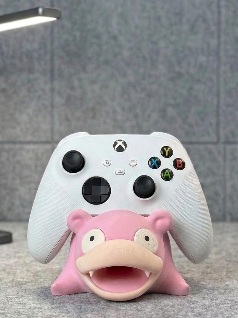 Pode incluir: Um controle Xbox branco repousa sobre um suporte rosa em forma de Slowpoke. O controle tem joysticks pretos e bot&otilde;es coloridos. O suporte &eacute; uma figura tridimensional detalhada do personagem Pok&eacute;mon.