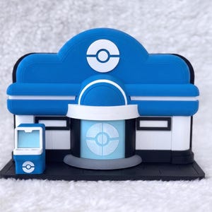 May include: A blue and white miniature building model featuring a central dome with a circular emblem, a light blue entrance door with a matching emblem, and a small blue arcade-style machine on the left side, all set on a black rectangular base.