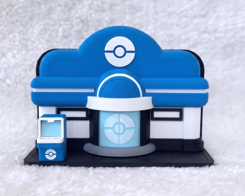 May include: A blue and white miniature building model featuring a central dome with a circular emblem, a light blue entrance door with a matching emblem, and a small blue arcade-style machine on the left side, all set on a black rectangular base.