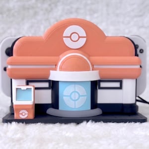 May include: A peach, white, and light blue plastic charging dock shaped like a building with a circular emblem on the roof and front door. The structure features a small matching kiosk on the left side and holds a white gaming console behind it.