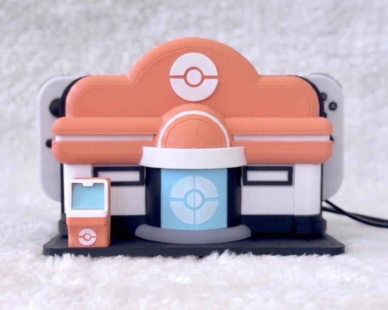 May include: A peach, white, and light blue plastic charging dock shaped like a building with a circular emblem on the roof and front door. The structure features a small matching kiosk on the left side and holds a white gaming console behind it.