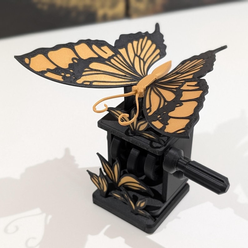 May include: A black and gold butterfly-shaped object with a crank. The butterfly has detailed wing patterns in gold and black. The base is a black box with a crank handle and decorative gold accents. The object is on a white surface.