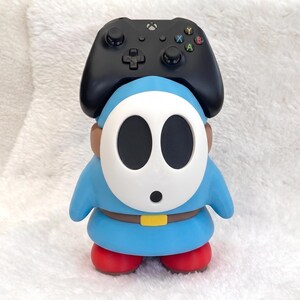 May include: A blue Shy Guy character figure with a white mask, brown belt, and red shoes, serving as a stand for a black Xbox wireless controller. The controller features a central Xbox logo, a directional pad, two analog sticks, and four colored buttons.