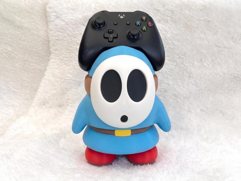 May include: A blue Shy Guy character figure with a white mask, brown belt, and red shoes, serving as a stand for a black Xbox wireless controller. The controller features a central Xbox logo, a directional pad, two analog sticks, and four colored buttons.