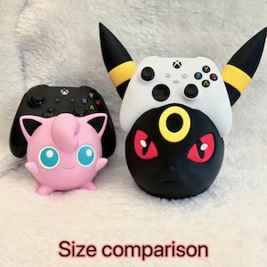 May include: A pink Jigglypuff figure holding a black Xbox controller and a black and yellow Umbreon figure holding a white Xbox controller. The text "Size comparison" is printed in red at the bottom of the image, showing two distinct gaming stands.
