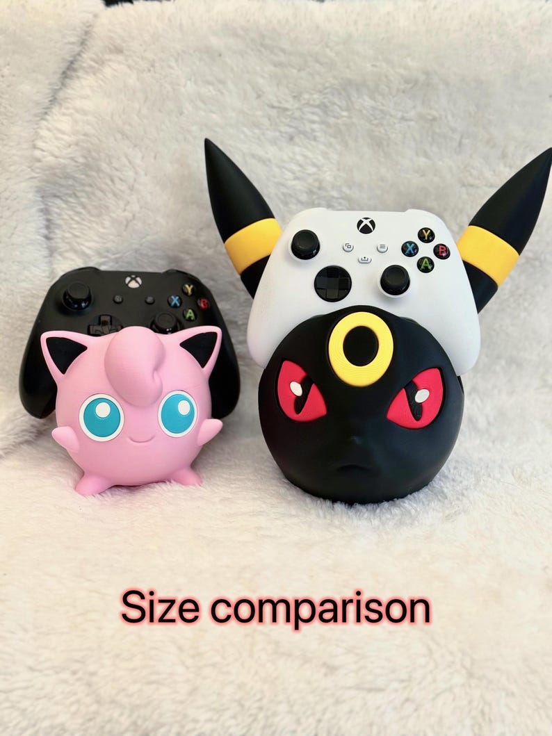 May include: A pink Jigglypuff figure holding a black Xbox controller and a black and yellow Umbreon figure holding a white Xbox controller. The text "Size comparison" is printed in red at the bottom of the image, showing two distinct gaming stands.