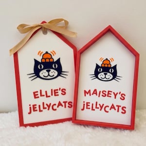 Puede incluir: Dos letreros de madera con forma de casa, marcos rojos y centros blancos. Cada letrero muestra una ilustraci&oacute;n de un gato negro con un sombrero naranja y el texto "ELLIE'S JELLYCATS" o "MAISEY'S JELLYCATS" en color rojo.