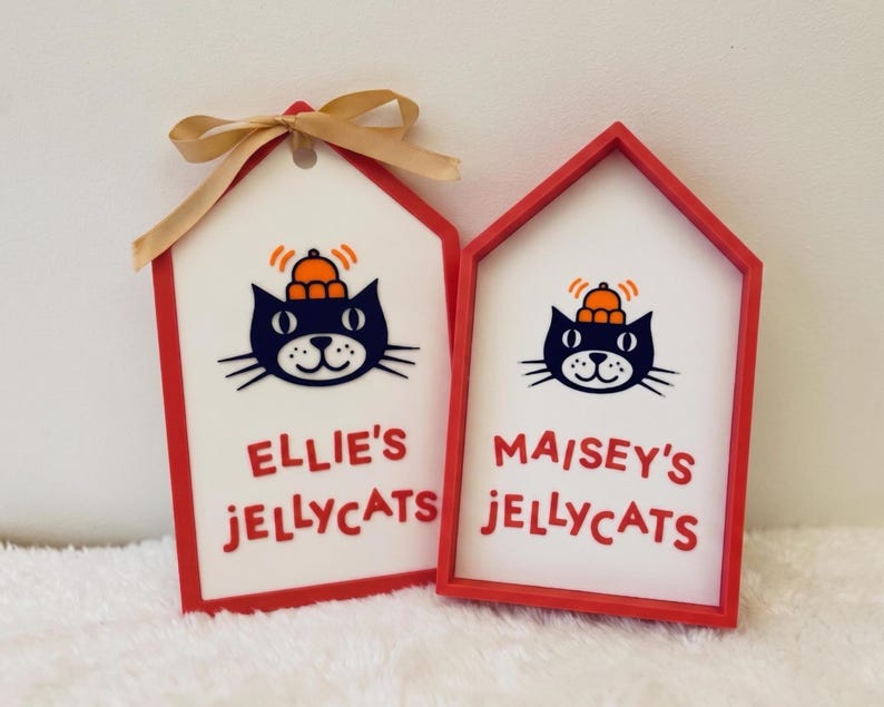 Puede incluir: Dos letreros de madera con forma de casa, marcos rojos y centros blancos. Cada letrero muestra una ilustraci&oacute;n de un gato negro con un sombrero naranja y el texto "ELLIE'S JELLYCATS" o "MAISEY'S JELLYCATS" en color rojo.