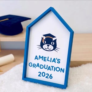 May include: A blue framed sign with the text "AMELIA'S GRADUATION 2026" and a cat wearing a graduation cap. A graduation cap and diploma are in the background. The sign is on a white surface.