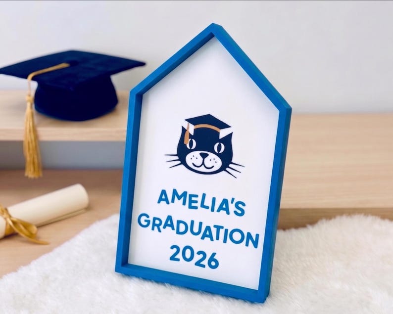 May include: A blue framed sign with the text "AMELIA'S GRADUATION 2026" and a cat wearing a graduation cap. A graduation cap and diploma are in the background. The sign is on a white surface.