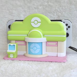 May include: A lime green and beige gaming console dock shaped like a building with a central blue door, a white circular emblem on the roof, and a small green arcade-style machine on a pink base, holding a white gaming controller with black buttons.