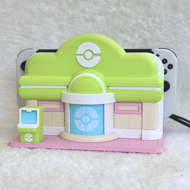 May include: A lime green and beige gaming console dock shaped like a building with a central blue door, a white circular emblem on the roof, and a small green arcade-style machine on a pink base, holding a white gaming controller with black buttons.
