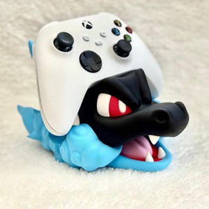 May include: A white wireless gaming controller resting on a black and light blue creature-shaped stand. The stand features a black head with red eyes, a wide open mouth with white teeth and a pink tongue, and a light blue body with textured details.