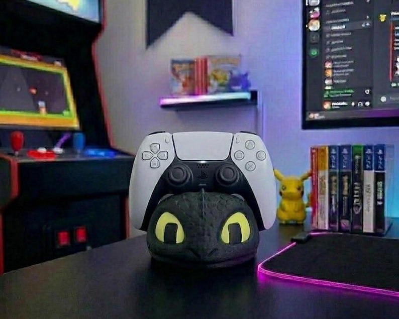 May include: A white and black PlayStation 5 controller sits atop a black dragon-shaped stand with yellow eyes. A yellow Pikachu figure and game cases are in the background. The scene is set on a black desk.