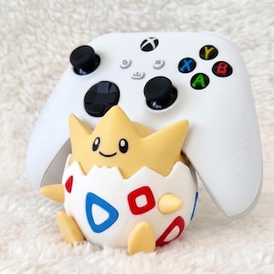 May include: A white wireless video game controller positioned behind a yellow and white character figurine. The figurine features a cracked egg shell design with red and blue triangle patterns, small yellow limbs, and a smiling face.