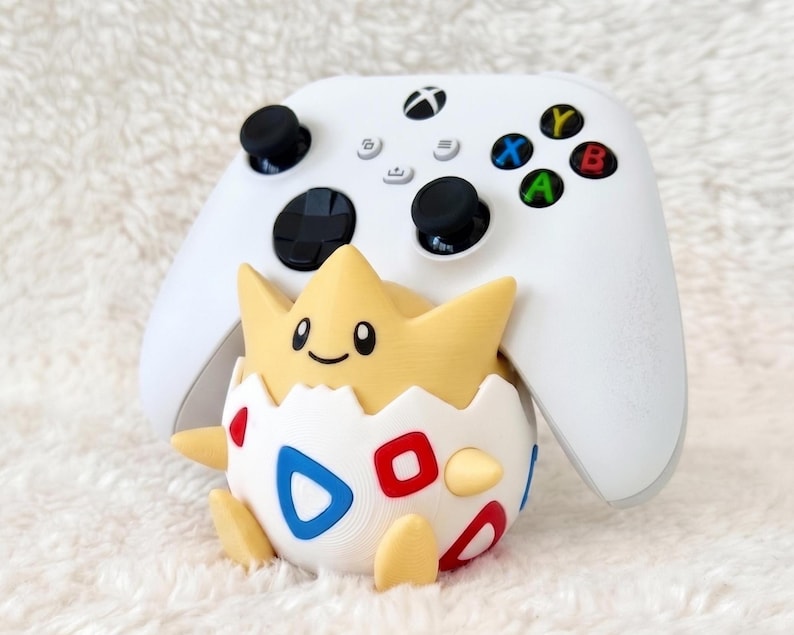 May include: A white wireless video game controller positioned behind a yellow and white character figurine. The figurine features a cracked egg shell design with red and blue triangle patterns, small yellow limbs, and a smiling face.