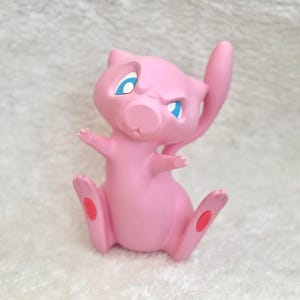 May include: A small, pink, feline-like creature figurine with large, pointed ears, a long, slender tail, and bright blue eyes. The character has a smooth, matte finish, small arms extended outward, and distinct red circular pads on its feet.