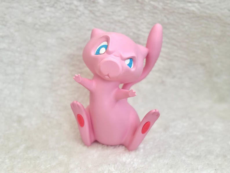May include: A small, pink, feline-like creature figurine with large, pointed ears, a long, slender tail, and bright blue eyes. The character has a smooth, matte finish, small arms extended outward, and distinct red circular pads on its feet.