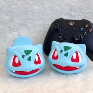 May include: Two light blue, bulbous, creature-shaped controllers or decorative covers featuring dark green spots on the forehead, large red eyes, and a wide mouth with visible white teeth, positioned in front of a black wireless gaming controller.
