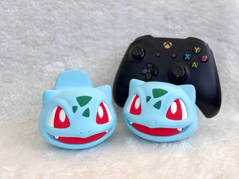 May include: Two light blue, bulbous, creature-shaped controllers or decorative covers featuring dark green spots on the forehead, large red eyes, and a wide mouth with visible white teeth, positioned in front of a black wireless gaming controller.