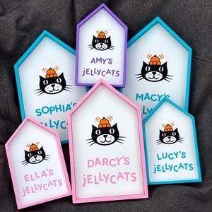 May include: A collection of six house-shaped signs with colorful frames. Each sign features a black cat wearing a pumpkin hat and the text "Amy's Jellycats", "Sophia's Lyc", "Macy's Ly", "Ella's Jellycats", "Darcy's Jellycats", and "Lucy's Jellycats".