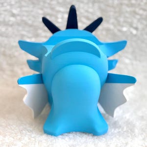 May include: A light blue figurine with a smooth matte finish, featuring a rounded central body, dark blue pointed crests on top, and white-tipped side fins. The object has a symmetrical structure with a wide base and distinct geometric shapes.
