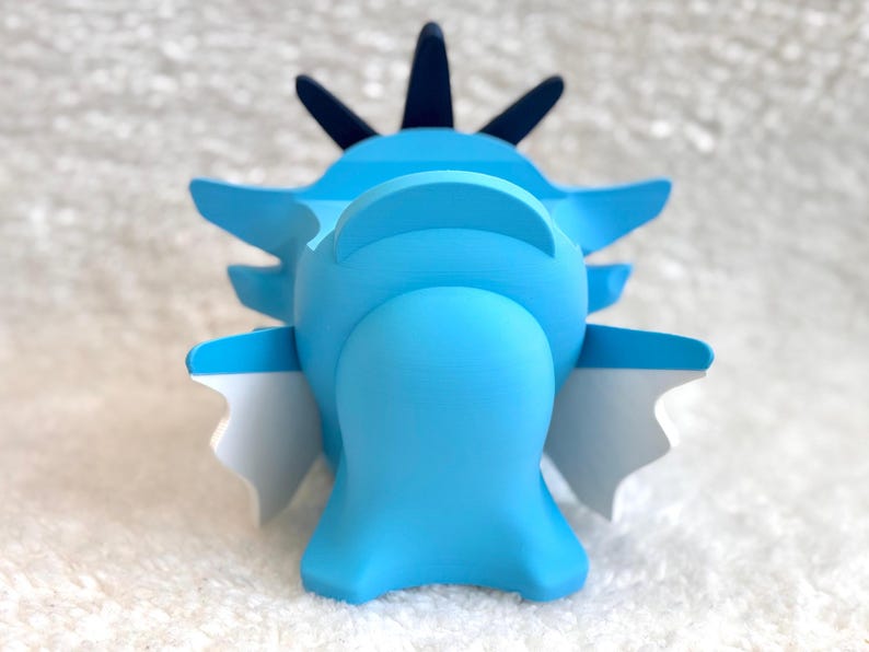 May include: A light blue figurine with a smooth matte finish, featuring a rounded central body, dark blue pointed crests on top, and white-tipped side fins. The object has a symmetrical structure with a wide base and distinct geometric shapes.