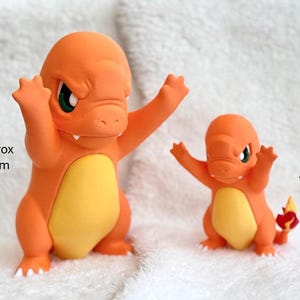 May include: Two orange and yellow Charmander figures with green eyes and raised arms. The larger figure measures approximately 9.06 inches in height, while the smaller figure measures approximately 5.91 inches in height, as indicated by the text.