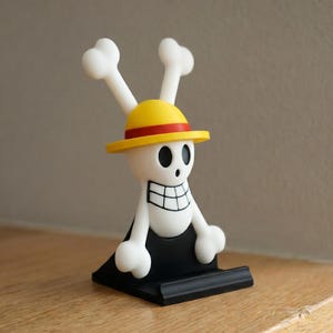 May include: A white and black figurine of a skull with a yellow hat and bone-shaped accents. The figurine has a black base and is set against a neutral background. The design is reminiscent of a popular anime series.