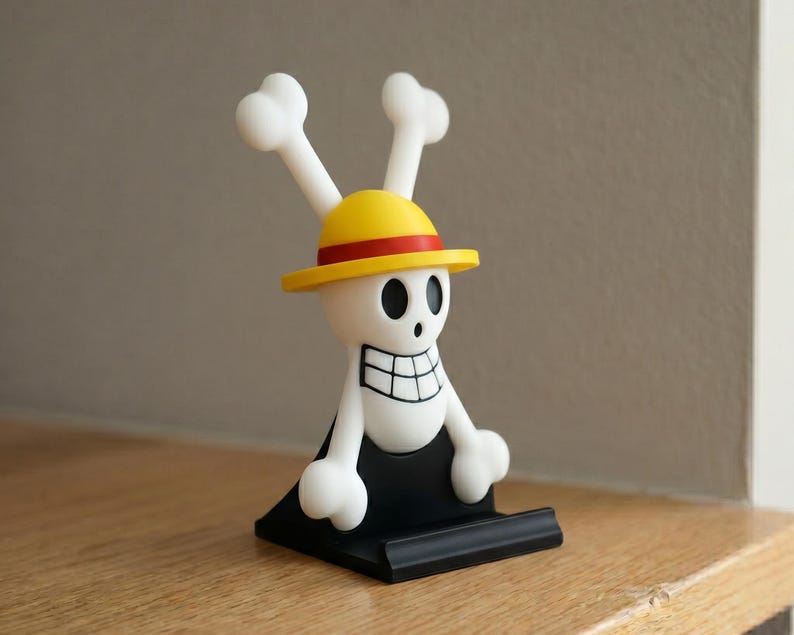 May include: A white and black figurine of a skull with a yellow hat and bone-shaped accents. The figurine has a black base and is set against a neutral background. The design is reminiscent of a popular anime series.