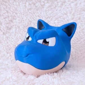 May include: A blue, stylized creature head figurine with pointed ears, dark inner ear details, and white eyes with black pupils. The figure features a smooth, matte finish, a prominent brow ridge, and a light beige lower jaw section, crafted in a solid form.