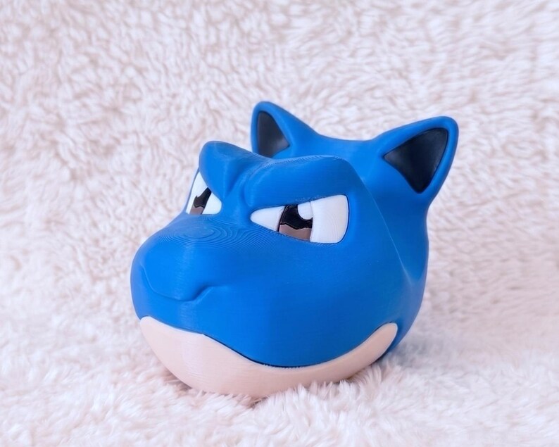 May include: A blue, stylized creature head figurine with pointed ears, dark inner ear details, and white eyes with black pupils. The figure features a smooth, matte finish, a prominent brow ridge, and a light beige lower jaw section, crafted in a solid form.