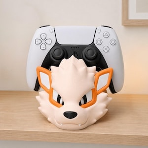Könnte beinhalten: Ein weißer PlayStation 5-Controller liegt auf einem hellpfirsichfarbenen Controllerhalter im Arkani-Design. Der Halter hat orangefarbene Akzente um Augen und Ohren, mit schwarzer Nase und Mund. Der Controller hat schwarze Knöpfe.