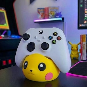 Puede incluir: Un mando blanco de Xbox descansa sobre una figura amarilla de Pikachu. El mando tiene joysticks negros y botones de colores. La figura de Pikachu tiene ojos negros y mejillas rosas. El fondo incluye elementos relacionados con videojuegos.